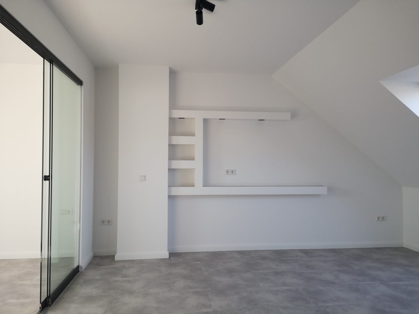 Immobilienmakler Münster Heller Raum mit schrägem Dach, weißer Wand und Regalen, grauem Boden und Glastür.